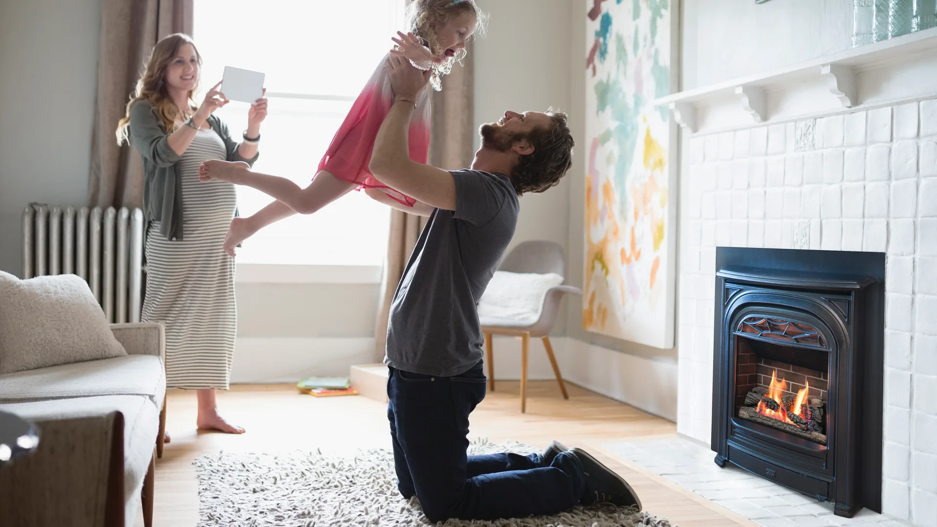 Father lifting his daughter in the air beside a gas fireplace insert with a white brick surround as the mother captures the moment